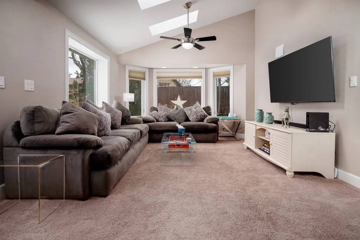 A wide, sunlit living room featuring a spacious sectional sofa oriented toward a wall-mounted large HD LED TV. Expansive windows flood the room with natural light, and cozy touches—like a decorative star motif and plush seating—create a welcoming, relaxed atmosphere ideal for gatherings or quiet moments.