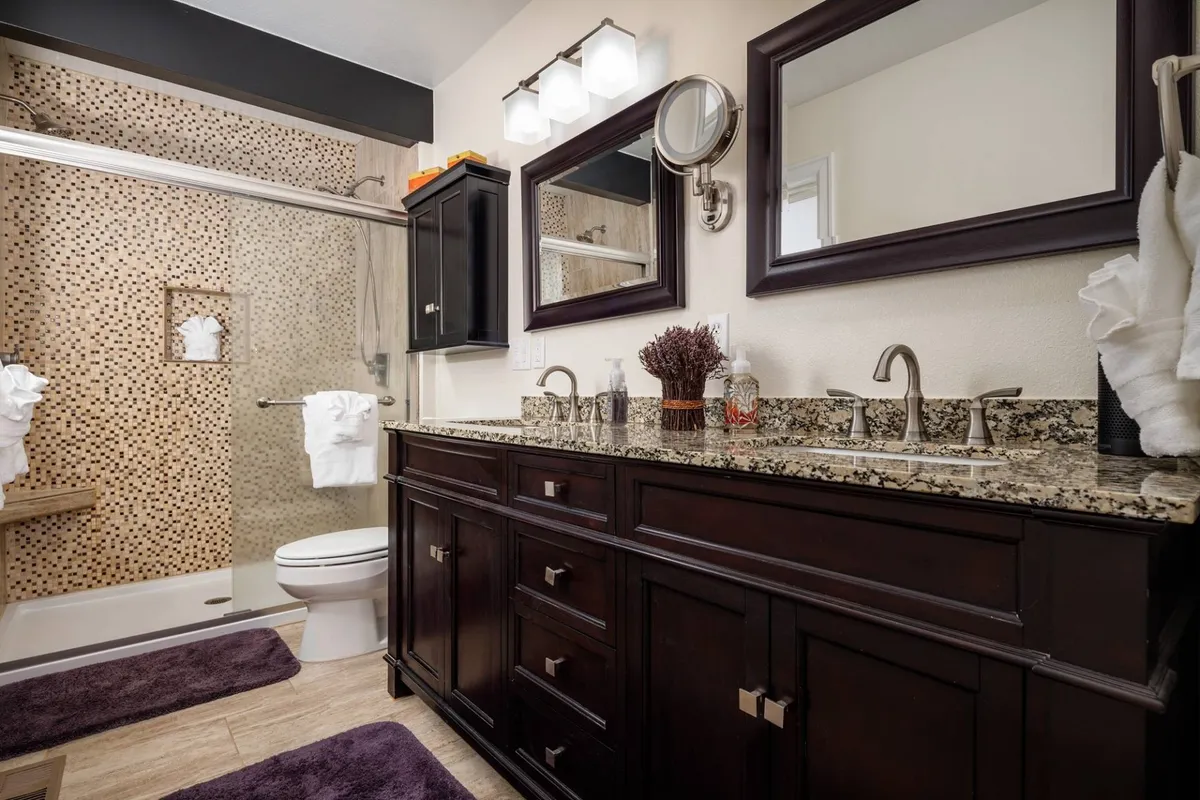 A refined bathroom featuring a dark wood vanity with granite countertop, framed mirrors, and a decorative shower wall. The space feels calm, clean, and organized, suitable for personal grooming or relaxing routines.