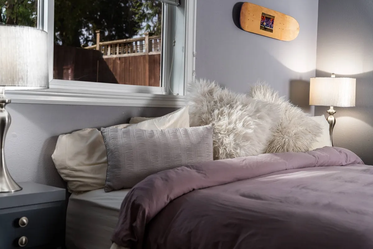 A serene bedroom scene showcases a neatly made bed with plush purple bedding and fluffy pillows, complemented by a soft table lamp and a playful skateboard wall decoration. The environment is calm and inviting, perfect for peaceful nights and restorative sleep.