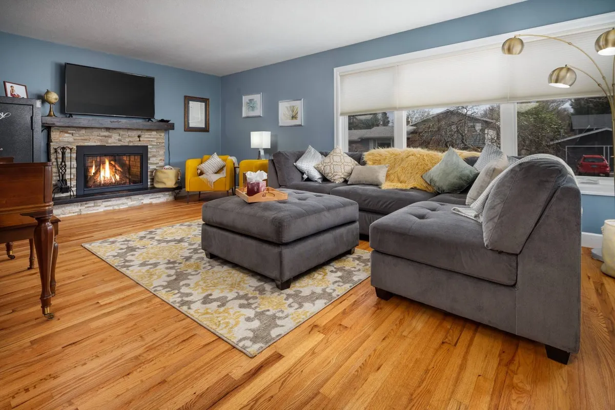 The image showcases a warm, inviting living room centered around a glowing fireplace, with a cozy sofa set and plush rug arranged for conversation or relaxation. Large windows let in natural light and offer a tranquil view of the outdoors, while tasteful artwork adds a touch of style.