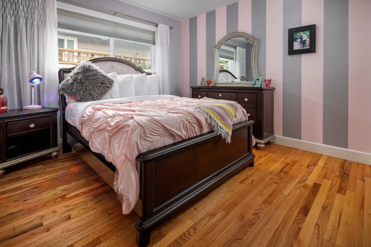 A cheerful bedroom with a bed dressed in a pink bedspread, a striped accent wall, a mirror, and a nightstand. The room feels playful yet comfortable, ideal for families or younger guests seeking a cozy retreat.