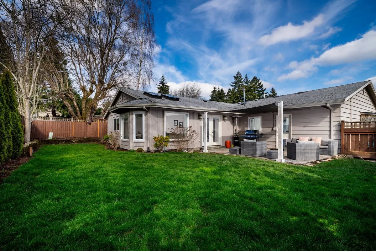 The photo captures the well-manicured backyard with a lush lawn, an attached patio area, and outdoor furniture. The yard is fully fenced, giving a clear impression of a secure space for children and pets to play. The mood is peaceful and inviting, with hints of both daytime and sunset enjoyment.