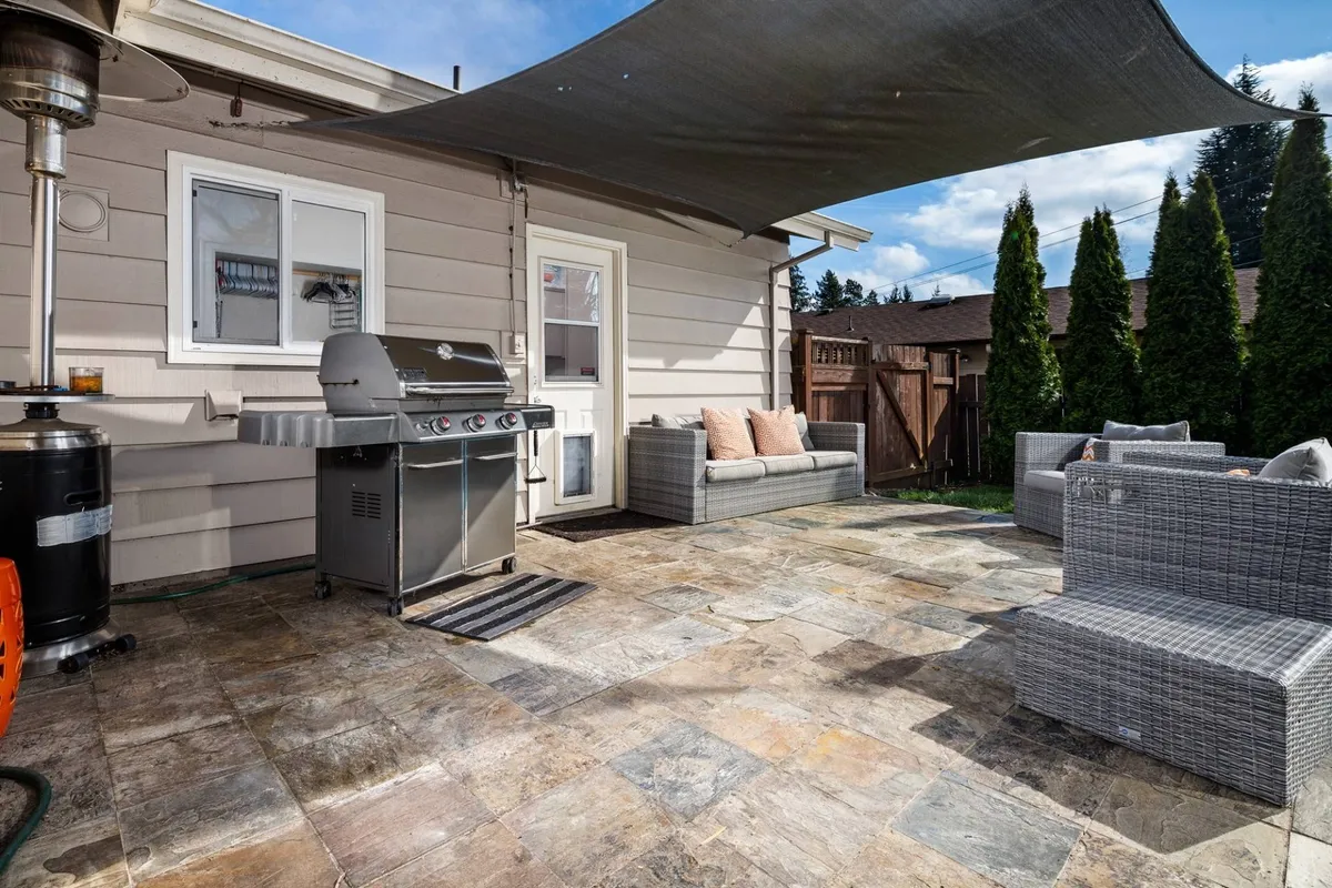 This image presents the sail-covered patio with comfortable outdoor seating and a BBQ grill as its focal points. The patio area is shaded by the sail overhead, with ample space for socializing, dining, or relaxing. The scene gives a sense of a welcoming, functional outdoor living area where families and friends can gather.