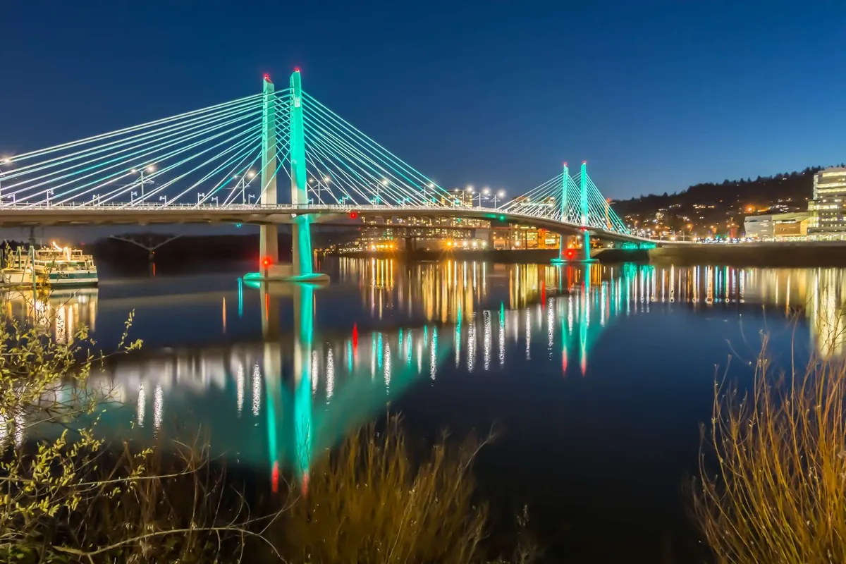 A picturesque view of a city bridge illuminated at night, with reflections dancing across the calm water and city lights glowing in the background. The scene captures an inviting evening atmosphere, hinting at the proximity to vibrant downtown life and local attractions.